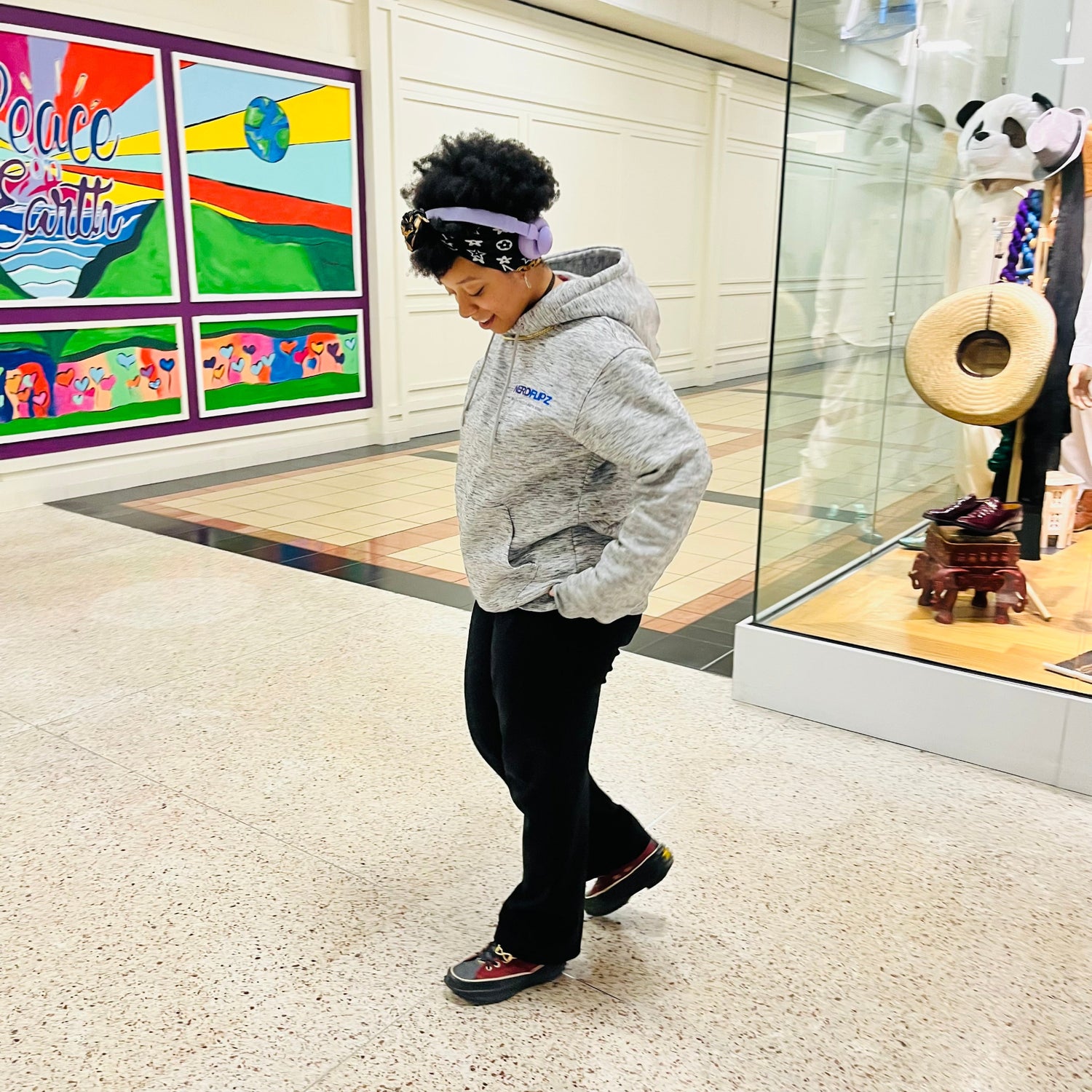 Person wearing a gray hoodie and black pants standing in a store with colorful artwork on the wall.